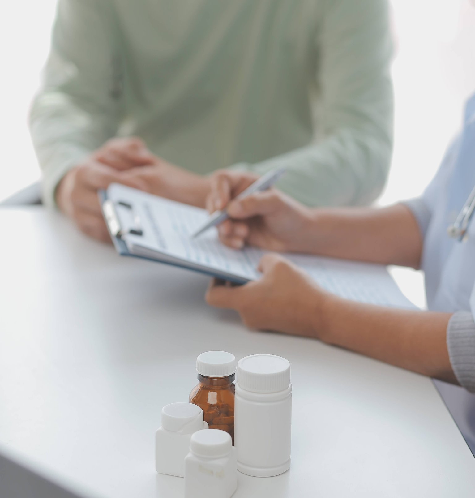 Two individuals sitting at a table, one writing on a clipboard, with a four different bottles of medication in the foreground.